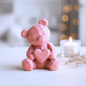 May include: A pink geometric teddy bear figurine with a heart-shaped chest. The bear is made of a series of connected triangular shapes. A lit candle and decorative sprigs are in the background.