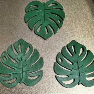 Puede incluir: Tres posavasos verdes con forma de hoja de Monstera. Los posavasos est&aacute;n hechos de un material s&oacute;lido y tienen un patr&oacute;n de hoja detallado con secciones recortadas. Los posavasos est&aacute;n dispuestos sobre una superficie texturizada de color claro.