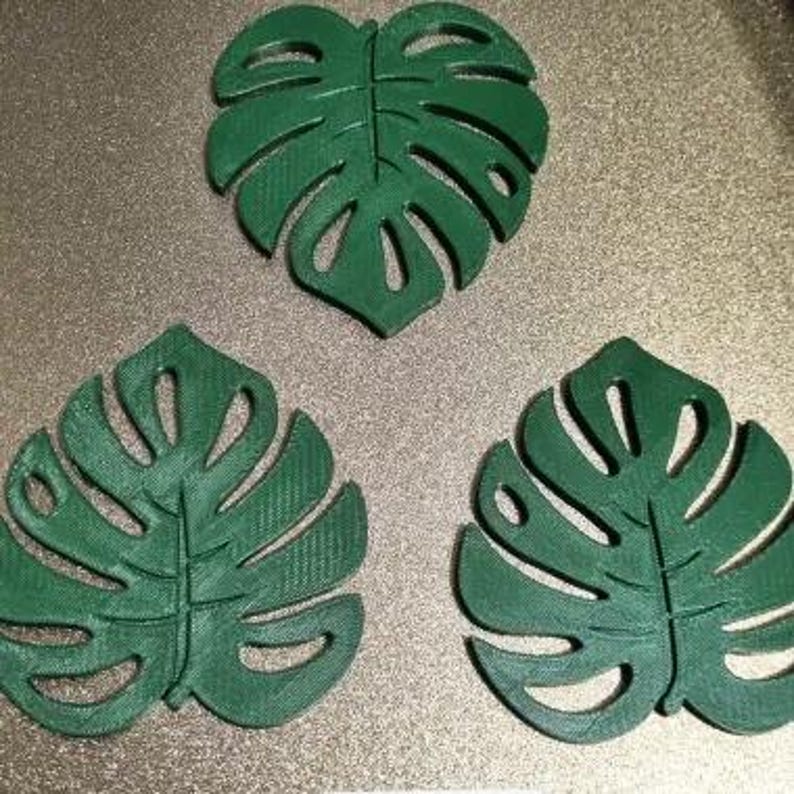 Puede incluir: Tres posavasos verdes con forma de hoja de Monstera. Los posavasos est&aacute;n hechos de un material s&oacute;lido y tienen un patr&oacute;n de hoja detallado con secciones recortadas. Los posavasos est&aacute;n dispuestos sobre una superficie texturizada de color claro.
