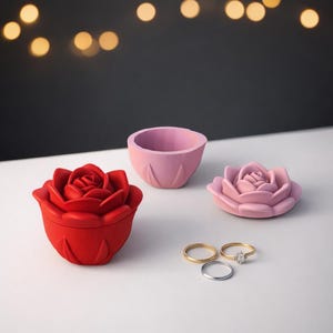 Könnte beinhalten: Eine rote rosenförmige Schachtel, eine rosa Schale und eine rosa rosenförmige Schale werden gezeigt. Zwei goldene Ringe und ein silberner Ring befinden sich neben der Schale. Der Hintergrund ist dunkel mit verschwommenen gelben Lichtern.