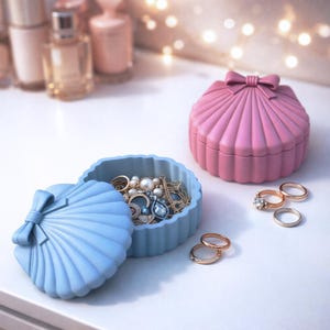 May include: A light blue seashell-shaped jewelry box with a bow on the lid, open to reveal necklaces, rings, and earrings. A pink seashell-shaped box with a bow is next to it. Several rings are scattered nearby. The boxes are on a white surface.