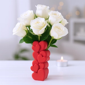Puede incluir: Un ramo de rosas blancas en un jarrón rojo en forma de corazón. El jarrón está hecho de corazones apilados. Las rosas tienen hojas verdes y están en plena floración. Una vela encendida está en el fondo.