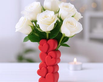 May include: A vase of white roses in a red heart-shaped vase. The vase is composed of stacked hearts. The roses have green leaves and are in full bloom. A lit candle is in the background.