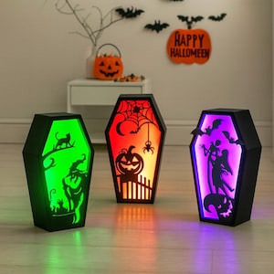 May include: Three black coffin-shaped Halloween decorations with illuminated interiors. One glows green with a witch silhouette, another red with a pumpkin and spider, and the last purple with a couple silhouette. The background includes a pumpkin and bats.