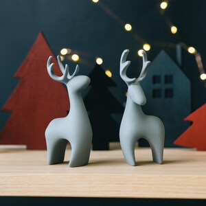 May include: Two light gray ceramic reindeer figurines with stylized antlers stand on a wooden surface. The background features red and black Christmas tree cutouts and a dark blue backdrop with string lights, creating a festive holiday scene.