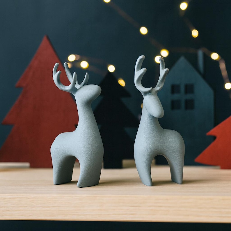 May include: Two light gray ceramic reindeer figurines with stylized antlers stand on a wooden surface. The background features red and black Christmas tree cutouts and a dark blue backdrop with string lights, creating a festive holiday scene.