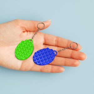 May include: Two oval-shaped keychains, one green and one blue, each with a textured, studded surface. The keychains are attached to silver-colored chains and key rings. The background is a solid light blue.