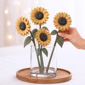 Op de afbeelding: Een heldere glazen vaas bevat verschillende kunstzonnebloemen. De zonnebloemen hebben gele bloemblaadjes, donkerbruine centra en groene stelen en bladeren. De vaas staat op een ronde houten dienblad.