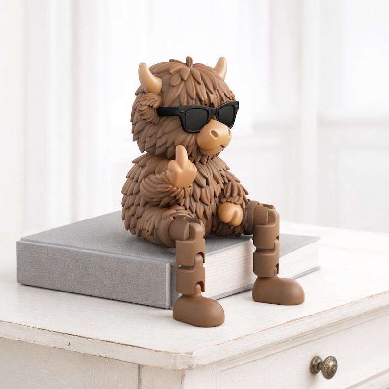 May include: A brown figurine of a Highland cow wearing black sunglasses and giving the middle finger. The cow is sitting on a gray book, with articulated legs. The figurine has a shaggy coat and small horns.