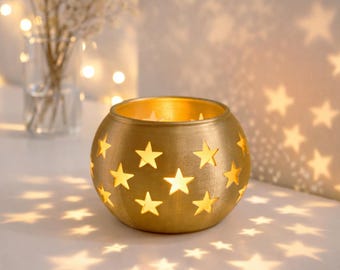 May include: A spherical gold-toned metal candle holder featuring numerous star-shaped cutouts. The interior is illuminated, casting star-shaped light patterns onto the surrounding surface. The exterior has a textured, metallic finish.