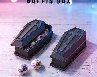 May include: Two black coffin-shaped boxes with a cross embossed on the lids. One box is open, revealing several round, eyeball-patterned candies inside. Two additional eyeball candies sit outside the boxes. The text COFFIN BOX is visible.
