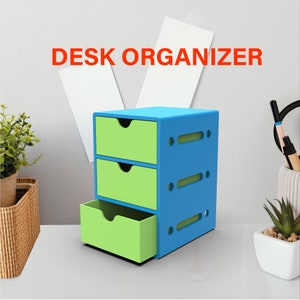 Puede incluir: Un organizador de escritorio azul y verde con tres cajones. El organizador tiene un dise&ntilde;o moderno con recortes en los lados. "DESK ORGANIZER" est&aacute; escrito en letras rojas en la pared detr&aacute;s del organizador.
