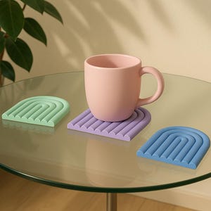 Puede incluir: Tres posavasos de silicona con forma de arco, uno verde, uno morado y uno azul. Una taza rosa está sobre el posavasos morado.