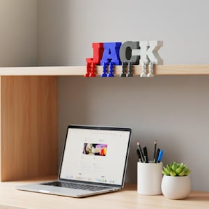 May include: A colorful, 3D-printed name sign spelling "JACK" in red, blue, gray, and white letters. Each letter has small legs, appearing to hang from a shelf. A laptop, pens, and a small succulent in a white pot are on the desk below.