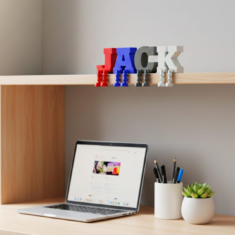 May include: A colorful, 3D-printed name sign spelling "JACK" in red, blue, gray, and white letters. Each letter has small legs, appearing to hang from a shelf. A laptop, pens, and a small succulent in a white pot are on the desk below.