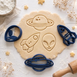 Puede incluir: Un juego de cortadores de galletas de plástico azul oscuro con formas temáticas del espacio, incluyendo un planeta con anillos, un cohete y una cabeza de alienígena, dispuestos sobre una masa de galletas circular con impresiones.