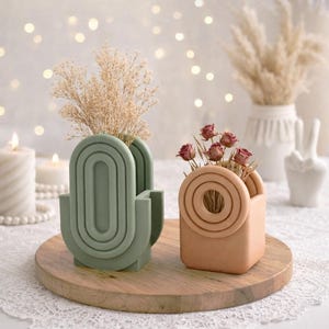 Peut inclure: Deux vases décoratifs, l'un vert sauge et l'autre pêche, contenant chacun des fleurs séchées. Le vase vert présente un design en arche, tandis que le vase pêche arbore un motif circulaire. Ils reposent sur un plateau en bois, créant une esthétique minimaliste.