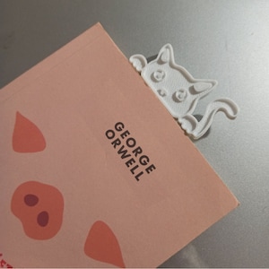 以下が含まれることがあります： 本から顔を出している猫の形をした白いプラスチック製のしおり。本の表紙はピンクで、豚のイラストと「GEORGE ORWELL」という文字が印刷されています。