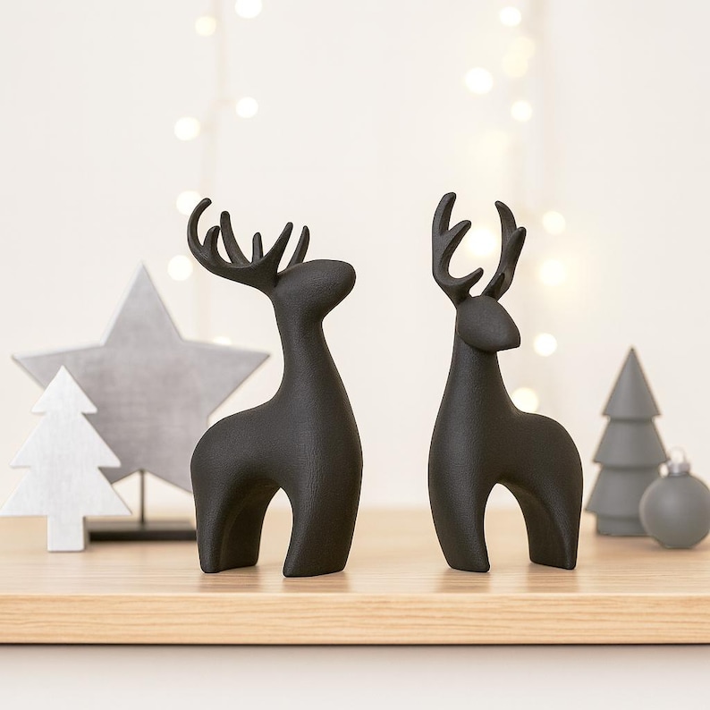 May include: Two matte black reindeer figurines with detailed antlers stand on a wooden surface. A gray star, a white tree, a gray tree, and a gray ornament are in the background, suggesting a holiday theme.