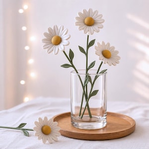 May include: A clear glass cylinder vase containing three artificial daisy flowers with white petals, yellow centers, and green stems with leaves. A fourth matching daisy flower lies horizontally on the surface next to the vase.