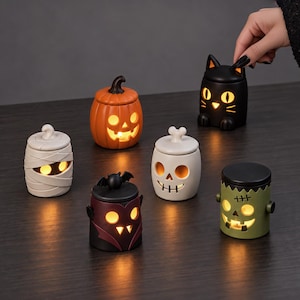 Pu&ograve; includere: Un set di sei portacandele in ceramica a tema Halloween raffiguranti una zucca, un gatto nero, una mummia, un vampiro con pipistrello, un teschio e un mostro verde, illuminati dall'interno per diffondere una luce calda dai volti intagliati.