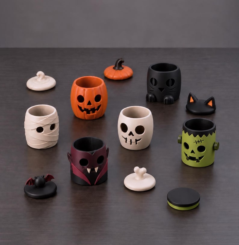 Pu&ograve; includere: Una collezione di piccoli contenitori cavi in ceramica con personaggi a tema Halloween, tra cui una zucca, una mummia, un vampiro, un teschio, un gatto nero e un mostro verde, ognuno con un coperchio abbinato o un elemento decorativo.