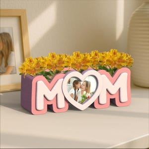 Puede incluir: Un macetero de cerámica rosa y blanco con forma de la palabra "MOM" con un marco de fotos en forma de corazón en el centro. El macetero está lleno de flores amarillas y una foto de una madre y su hija.