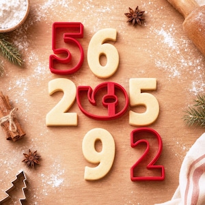 May include: A collection of red plastic number cookie cutters and baked light beige number cookies arranged on a wooden surface. The set includes the numbers five, six, two, nine, and a stylized zero, alongside star anise and flour.