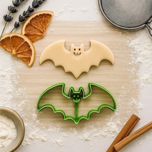 May include: A beige bat-shaped cookie and a green bat-shaped cookie cutter are displayed on a light wooden surface dusted with flour. The cookie has a smiling face with red eyes. Dried orange slices, lavender, and cinnamon sticks are also present.