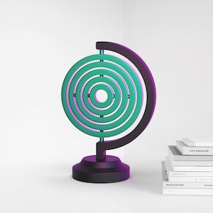 Puede incluir: Un soporte de globo de metal negro y morado con un dise&ntilde;o de c&iacute;rculos conc&eacute;ntricos verde y morado. El globo est&aacute; sobre una superficie blanca.