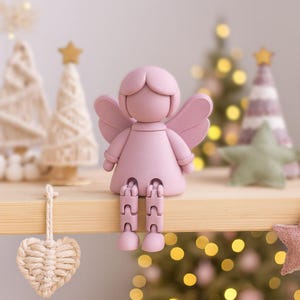 Puede incluir: Una figura de ángel rosa con piernas y alas articuladas está sentada en una estantería de madera. El ángel está frente a árboles de Navidad borrosos y un adorno en forma de estrella. La figura es un objeto decorativo.