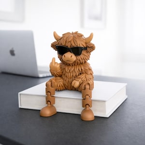 May include: A brown figurine of a Highland cow wearing black sunglasses, giving the middle finger. The cow is sitting on a white book with articulated legs. The figurine is on a dark surface, with a laptop in the background.