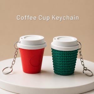Puede incluir: Dos llaveros de taza de café con tapas blancas y anillas de metal plateado. Una taza es de color rojo sólido con una superficie lisa, mientras que la otra es de color verde oscuro con un exterior texturizado y estriado. El texto Coffee Cup Keychain es visible.