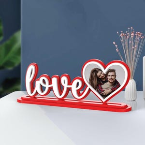 Peut inclure: Un panneau en bois rouge 'love' avec un cadre en forme de cœur blanc pour une photo. Le cadre en forme de cœur contient une photo d'un couple.