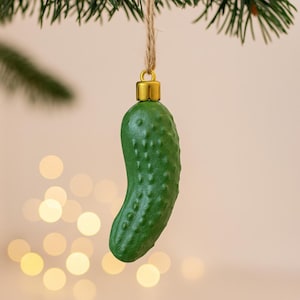 May include: A green pickle-shaped Christmas ornament with a gold-colored cap, hanging from a twine string. The ornament has a textured surface and is suspended against a blurred background with bokeh lights.