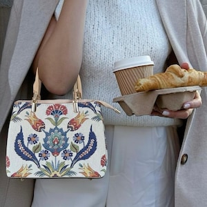 Może przedstawiać: Kremowa torebka z kwiatowym wzorem w kolorze czerwonym, niebieskim i żółtym. Torebka ma prostokątny kształt z górnym uchwytem i zapięciem na zamek błyskawiczny. Na zdjęciu widać również filiżankę kawy i rogalik.