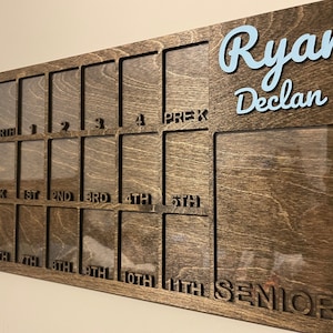 May include: A wooden photo display board with slots for pictures. The board has the names "Ryan Declan" at the top and the grades "Birth, 1st, 2nd, 3rd, 4th, 5th, 6th, 7th, 8th, 9th, 10th, 11th, Senior" printed below the slots.