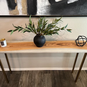 May include: A white and brown wood console table with gold metal legs. The table is decorated with a black vase with green leafy branches, a candle in a glass jar, and a black metal orb.