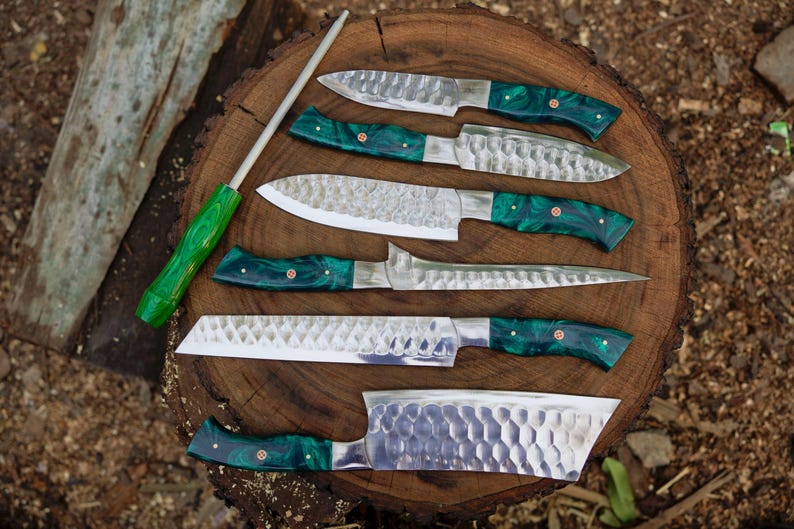 May include: A set of seven kitchen knives with green handles and a hammered finish. The knives are arranged on a wooden surface with a sharpening steel.