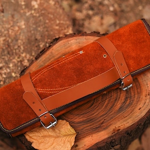 May include: A brown leather knife roll with a strap and buckle closure. The roll is lying on a wooden surface.