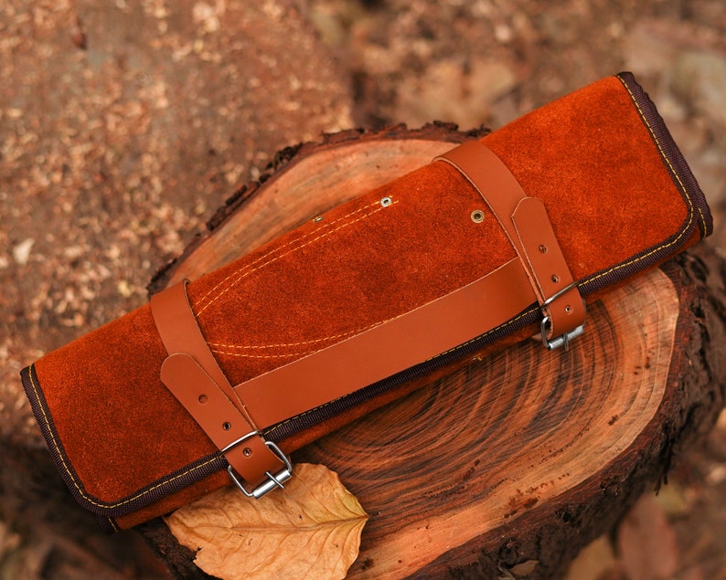 May include: A brown leather knife roll with a strap and buckle closure. The roll is lying on a wooden surface.