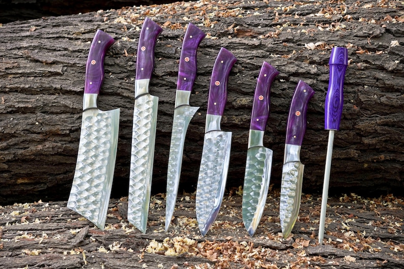 May include: A set of seven kitchen knives with purple handles and a steel sharpening steel. The knives have a hammered finish and are arranged on a wooden surface.