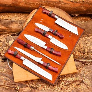 May include: A brown leather knife roll with six stainless steel knives and a sharpening steel. The knives have red and black handles.