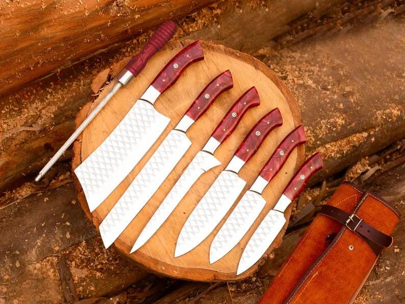 May include: Set of seven kitchen knives with red wood handles and a matching leather roll. The knives have a diamond pattern on the blades.
