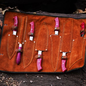 May include: A brown leather knife roll with six slots for knives. The knives have pink handles and are arranged in the roll.