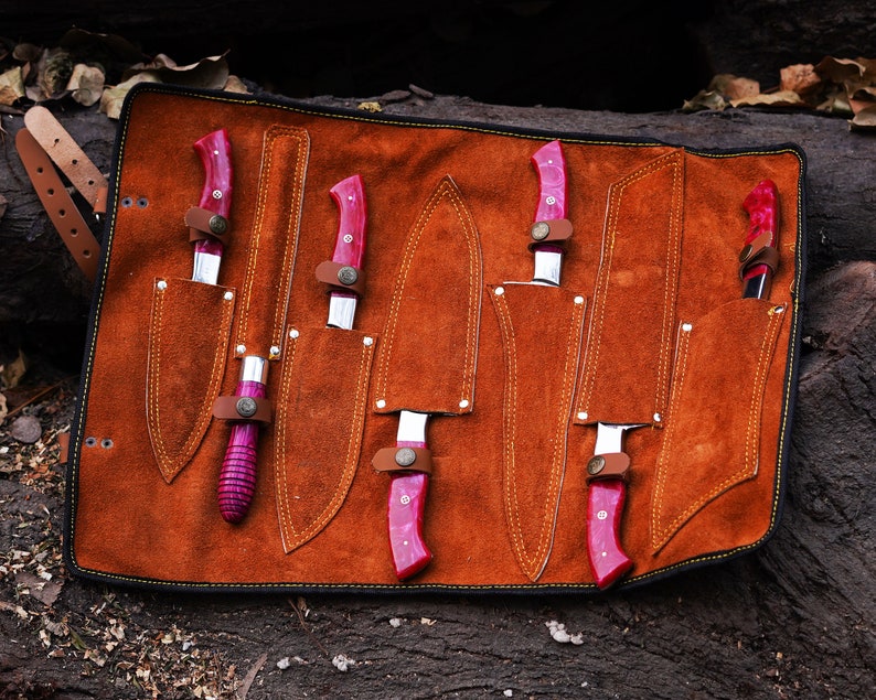 May include: A brown leather knife roll with six slots for knives. The knives have pink handles and are arranged in the roll.