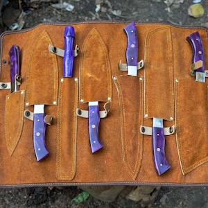 May include: A brown leather knife roll with six slots for knives. The roll is open and shows five knives with purple handles. The knives are all different sizes and shapes.