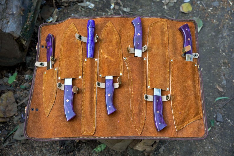 May include: A brown leather knife roll with six slots for knives. The roll is open and shows five knives with purple handles. The knives are all different sizes and shapes.
