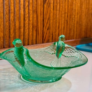 May include: A translucent light green glass bowl featuring two molded bird figures perched on opposite sides. The exterior surface of the bowl displays a textured, wavy pattern that mimics the appearance of a nest or woven material.