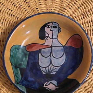 May include: A ceramic bowl with a stylized painting of a person in blue, green, red, and yellow on a yellow background.